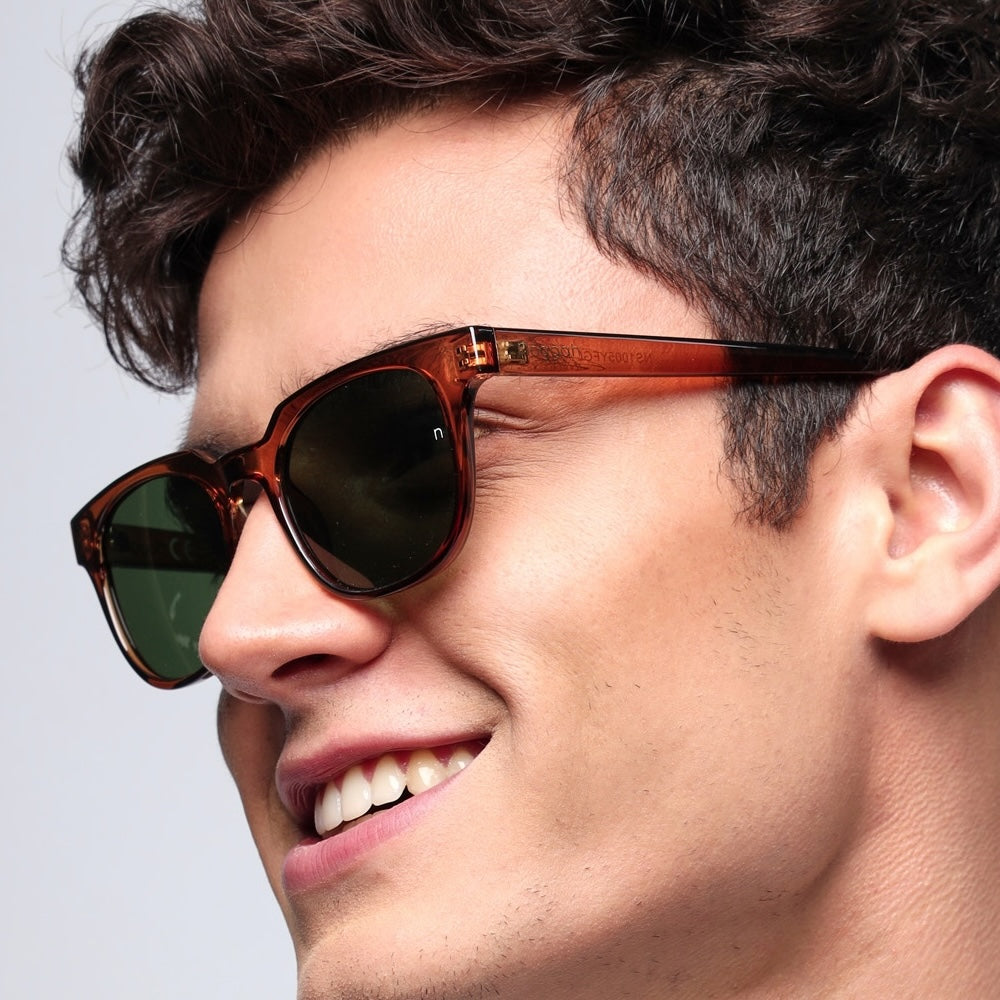 Model wearing Aura Desert Flame sunglasses (NS1005YFGL) with warm brown polycarbonate frame and scratch-resistant glass lenses under golden sunlight